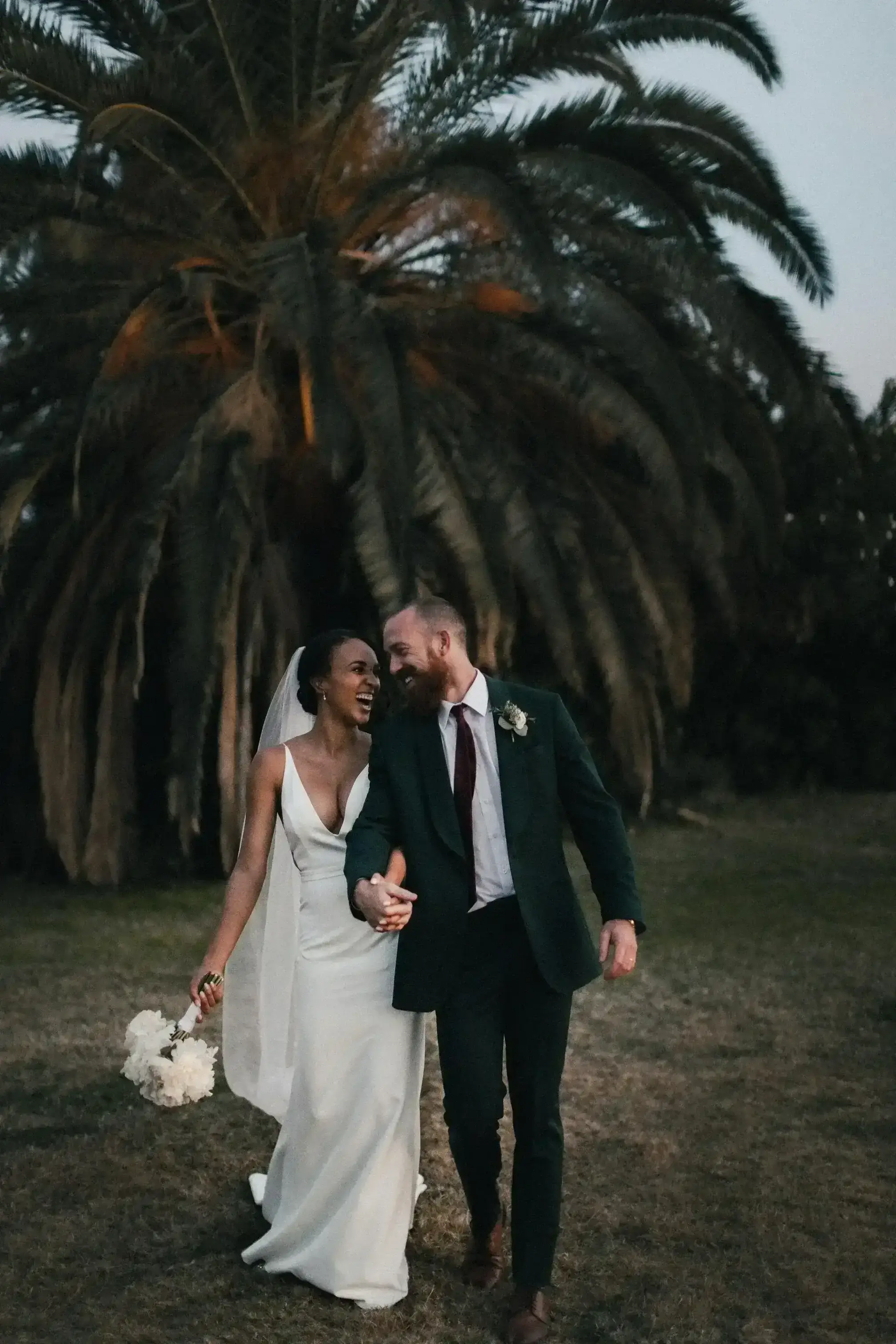 A woman in a wedding dress and her husband in a green suit are laughing under the palm trees.